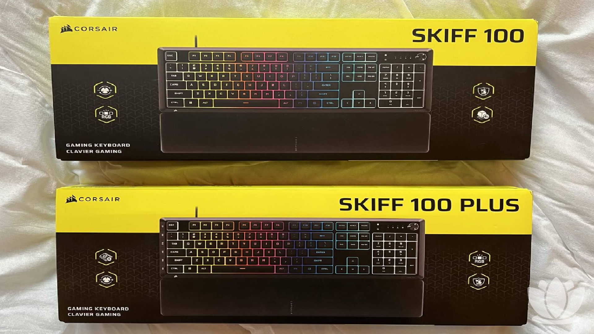 Corsair Skiff 100 and Skiff 100 Plus keyboards shown in their box on a white bedsheet.